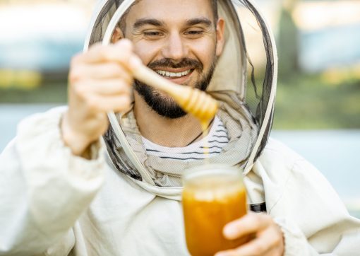 Portrait of a handsome beekeper in protective uniform standing with honey, tasting fresh product on the apiary outdoors